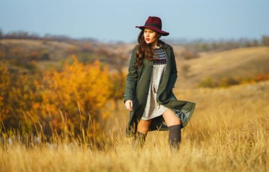 Sonbahar parkında şapkalı zarif bir kadın yürüyor. Çayırda sonbahar havasının tadını çıkaran şık bir kadın. Sonbahar modası. Dinlenme, rahatlama, yaşam tarzı konsepti.