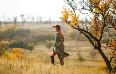 Sonbahar parkında şapkalı zarif bir kadın yürüyor. Çayırda sonbahar havasının tadını çıkaran şık bir kadın. Sonbahar modası. Dinlenme, rahatlama, yaşam tarzı konsepti.