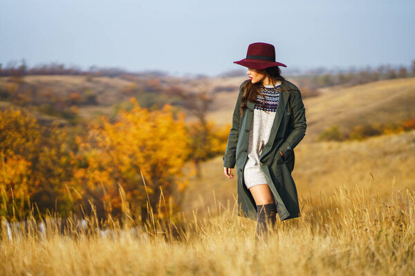 Elegant woman in coat with hat walks in autumn park. Stylish woman enjoying autumn weather in the meadow. Autumn fashion. Rest, relaxation, lifestyle concept.