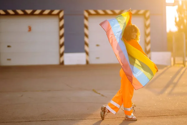 Young African American lesbian woman with LGBT rainbow flag in the street at sunset. Stylish woman with curly hair in an orange suit posing with lgbt pride flag.