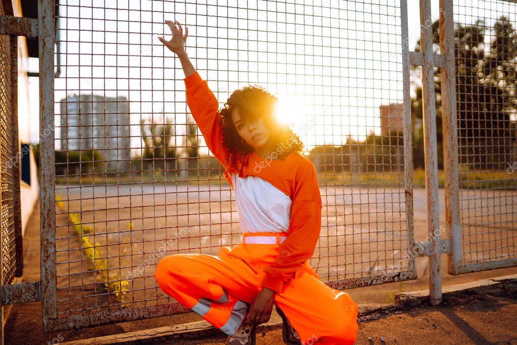 Young African American woman - dancer dancing in the street at sunset. Stylish woman with curly hair in an orange suit  showing some moves. Sport, dancing and urban culture concept.