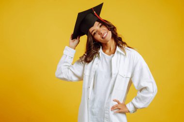 Mezuniyet şapkalı, sarı arka planda poz veren mezun bir kadın. Eğitim, eğitim, üniversite, üniversite, mezuniyet konsepti.