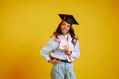 Mezuniyet şapkalı, sarı arka planda poz veren mezun bir kadın. Eğitim, eğitim, üniversite, üniversite, mezuniyet konsepti.