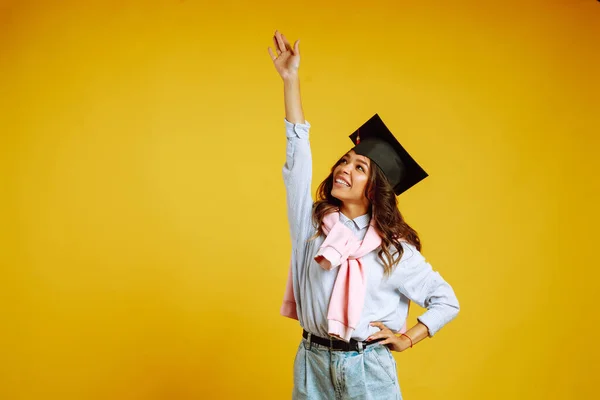 Graduate woman in a graduation hat on her head posing on a yellow background. Study, education, university, college, graduate concept.