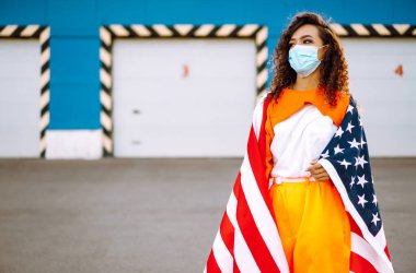Amerikan bayrağıyla poz veren tıbbi maskeli Afro-Amerikalı kadın. Karantina sırasındaki kıvırcık kadın. Salgının yayılmasını önleme kavramı. Covid 2019.
