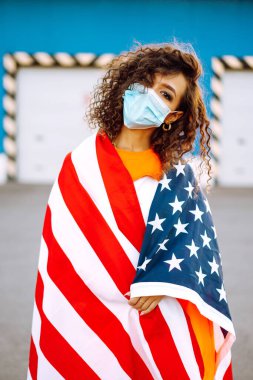 Amerikan bayrağıyla poz veren tıbbi maskeli Afro-Amerikalı kadın. Karantina sırasındaki kıvırcık kadın. Salgının yayılmasını önleme kavramı. Covid 2019.