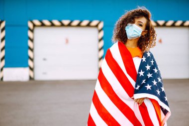 Amerikan bayrağıyla poz veren tıbbi maskeli Afro-Amerikalı kadın. Karantina sırasındaki kıvırcık kadın. Salgının yayılmasını önleme kavramı. Covid 2019.