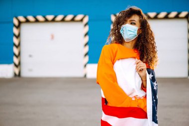 Amerikan bayrağıyla poz veren tıbbi maskeli Afro-Amerikalı kadın. Karantina sırasındaki kıvırcık kadın. Salgının yayılmasını önleme kavramı. Covid 2019.