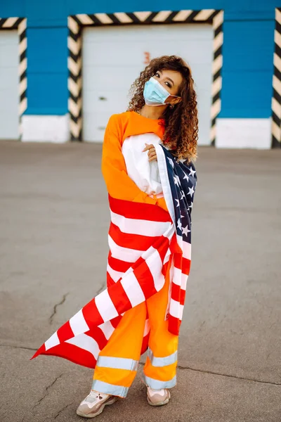 African American woman with medical face mask posing with american flag. Curly woman during quarantine. The concept of preventing the spread of the epidemic. Covid-2019.