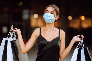 Yüzünde koruyucu steril tıbbi maske takmış alışveriş torbalarıyla gezen genç bir kadın. Satın alma, kara cuma, satış konsepti. Kapsam- 2019.