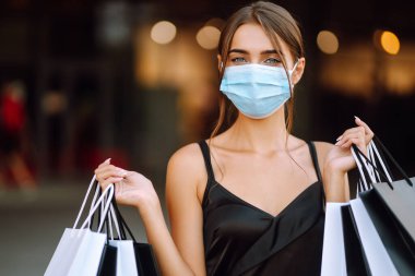 Yüzünde koruyucu steril tıbbi maske takmış alışveriş torbalarıyla gezen genç bir kadın. Satın alma, kara cuma, satış konsepti. Kapsam- 2019.
