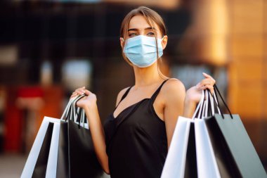 Alışveriş merkezinin yakınında alışveriş torbaları olan koruyucu steril tıbbi maskeli güzel bir kadın. Satın alma, kara cuma, satış konsepti. Kapsam- 2019.