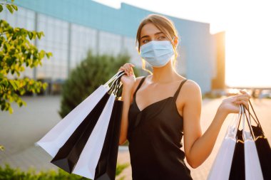 Güneş batarken alışveriş torbalarıyla koruyucu tıbbi maske takan şık bir kadın. Satın alma, kara cuma, satış konsepti. Kapsam- 2019.