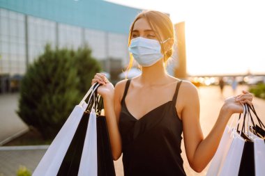 Güneş batarken alışveriş torbalarıyla koruyucu tıbbi maske takan şık bir kadın. Satın alma, kara cuma, satış konsepti. Kapsam- 2019.