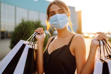 Güneş batarken alışveriş torbalarıyla koruyucu tıbbi maske takan şık bir kadın. Satın alma, kara cuma, satış konsepti. Kapsam- 2019.