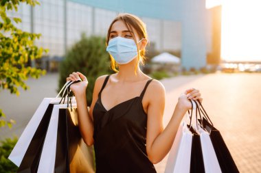 Güneş batarken alışveriş torbalarıyla koruyucu tıbbi maske takan şık bir kadın. Satın alma, kara cuma, satış konsepti. Kapsam- 2019.