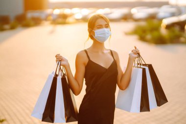 Güneş batarken alışveriş torbalarıyla koruyucu tıbbi maske takan şık bir kadın. Satın alma, kara cuma, satış konsepti. Kapsam- 2019.