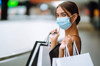 Alışveriş yapan bir kadın. Alışveriş merkezinde, yüzünde alışveriş torbalarıyla koruyucu steril tıbbi maske takan genç bir kadın. Satın almalar, kara cuma, indirimler, satış konsepti. Covid 2019.
