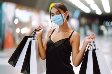 Alışveriş yapan bir kadın. Alışveriş merkezinde, yüzünde alışveriş torbalarıyla koruyucu steril tıbbi maske takan genç bir kadın. Satın almalar, kara cuma, indirimler, satış konsepti. Covid 2019.