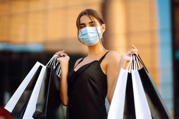 Beautiful woman in protective sterile medical mask with shopping bags near shopping center. Purchases, black friday, sale concept. Covid- 2019.