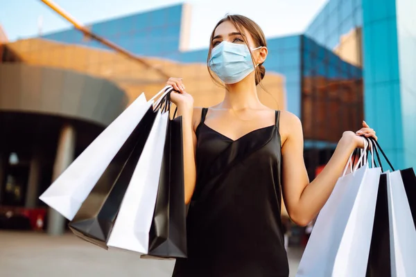 Beautiful woman in protective sterile medical mask with shopping bags near shopping center. Purchases, black friday, sale concept. Covid- 2019.