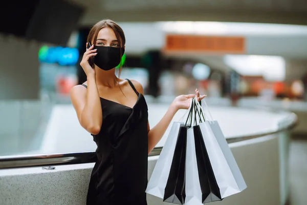 ショッピングバッグがモール内の携帯電話で話して保護黒医療マスクでファッショナブルな若い女性 黒の金曜日 販売の概念 Covid 2019 — ストック写真