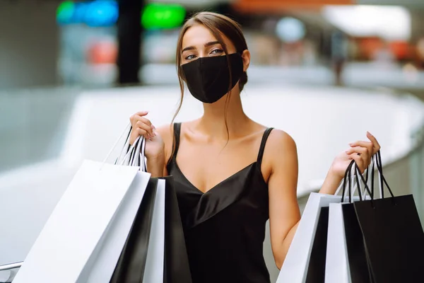 Young woman in protective black medical mask on her face with shopping bags in the mall. Purchases, black friday, discounts, sale concept. Covid-2019.