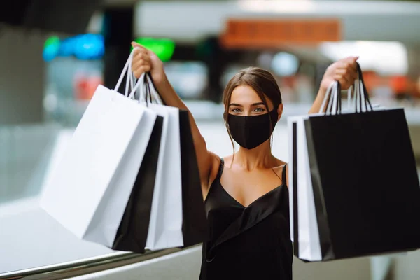 Young woman in protective black medical mask on her face with shopping bags in the mall. Purchases, black friday, discounts, sale concept. Covid-2019.