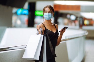 Alışveriş merkezinde alışveriş torbalarıyla koruyucu tıbbi maske takan şık bir kadın. Siyah elbiseli genç bir kadın cep telefonuyla konuşuyor. Satın alma, kara cuma, satış konsepti. Kapsam- 2019.