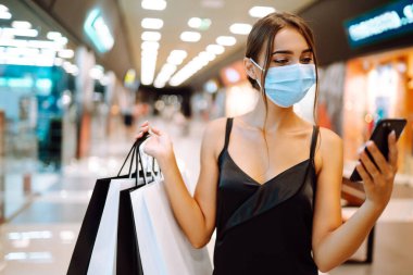 Alışveriş merkezinde alışveriş torbalarıyla koruyucu tıbbi maske takan şık bir kadın. Siyah elbiseli genç bir kadın cep telefonuyla konuşuyor. Satın alma, kara cuma, satış konsepti. Kapsam- 2019.