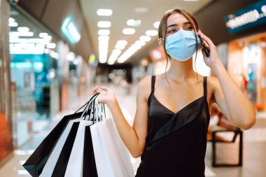 Alışveriş merkezinde alışveriş torbalarıyla koruyucu tıbbi maske takan şık bir kadın. Siyah elbiseli genç bir kadın cep telefonuyla konuşuyor. Satın alma, kara cuma, satış konsepti. Kapsam- 2019.