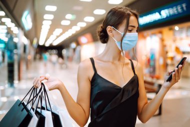 Alışveriş merkezinde alışveriş torbalarıyla koruyucu tıbbi maske takan şık bir kadın. Siyah elbiseli genç bir kadın cep telefonuyla konuşuyor. Satın alma, kara cuma, satış konsepti. Kapsam- 2019.