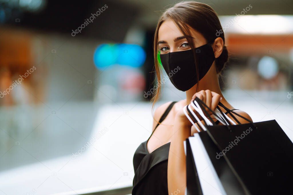 Young woman in protective black medical mask on her face with shopping bags in the mall. Purchases, black friday, discounts, sale concept. Covid-2019.