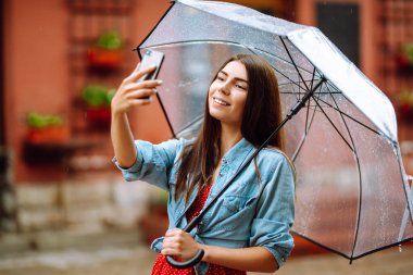 Genç bir kadın şehir caddesinde şeffaf bir şemsiyeyle selfie çekiyor. Şemsiye altında telefon kullanan gülümseyen kadın. Sonbahar konsepti.