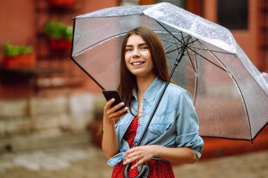 Şemsiye altında telefon kullanan gülümseyen bir kadının portresi. Şehir caddesinde poz veren şeffaf şemsiyeli genç bir kadın. Sonbahar konsepti.