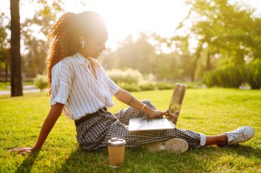 Laptop ve kahveyle yeşil çimlerde oturan genç bir kadın. Bilgisayarı ve kablosuz kulaklığı video görüşmesi için kullanan gülümseyen kadın. Gün batımı ışığı. Yaşam tarzı kavramı.