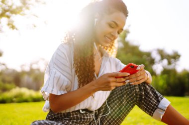 Parktaki yeşil çimlerin üzerinde oturan güzel genç bir kadının portresi. Şirin bir kadın cep telefonundan hoş bir mesaj okuyor ve müzik dinliyor..