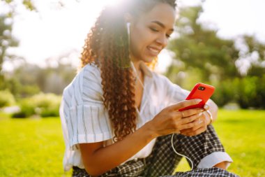 Parktaki yeşil çimlerin üzerinde oturan güzel genç bir kadının portresi. Şirin bir kadın cep telefonundan hoş bir mesaj okuyor ve müzik dinliyor..