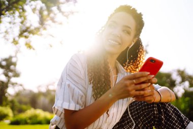 Parktaki yeşil çimlerin üzerinde oturan güzel genç bir kadının portresi. Şirin bir kadın cep telefonundan hoş bir mesaj okuyor ve müzik dinliyor..