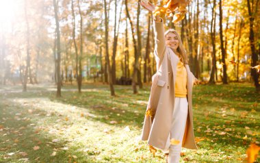 Mutlu genç kadın sonbahar parkında eğleniyor ve sarı yapraklarla oynuyor. Sonbahar moda konsepti.