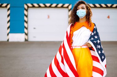 Koronavirüse karşı koruyucu bir maske takan ve elinde ABD bayrağı tutan Afro-Amerikalı bir kadın..