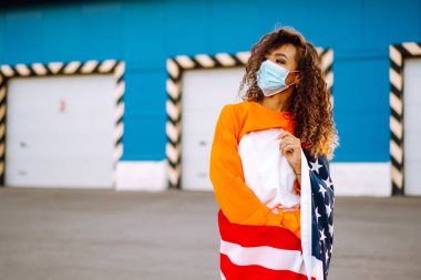 Koronavirüse karşı koruyucu bir maske takan ve elinde ABD bayrağı tutan Afro-Amerikalı bir kadın..