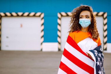 Koronavirüse karşı koruyucu bir maske takan ve elinde ABD bayrağı tutan Afro-Amerikalı bir kadın..