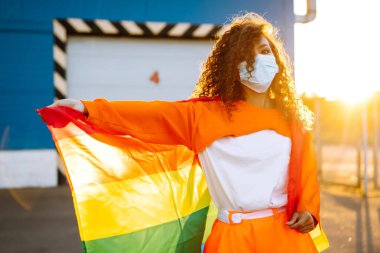 Koronavirüs salgını karantinası sırasında lgbt gurur bayrağı taşıyan koruyucu bir maske takan Afrikalı lezbiyen kadın. Covid 2019. LGBT transseksüel gökkuşağı konsepti.