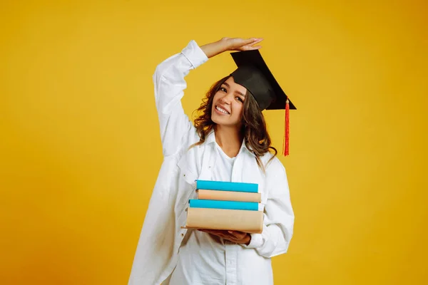 Smiling young woman in graduation hat  with books. Study, education, university, college, graduate concept on yellow banner.