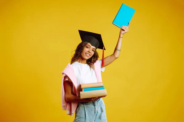 Smiling young woman in graduation hat  with books. Study, education, university, college, graduate concept on yellow banner.