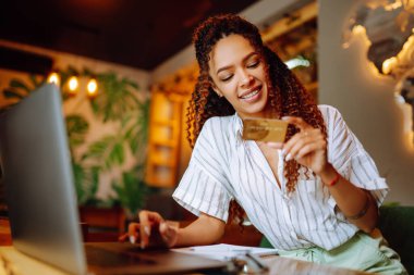 Kredi kartı tutan ve dizüstü bilgisayar kullanan genç bir kadın. Afro-Amerikalı bir kadın kafede oturmuş online alışveriş yapıyor. Çevrimiçi alışveriş, e-ticaret, internet bankacılığı, harcama.