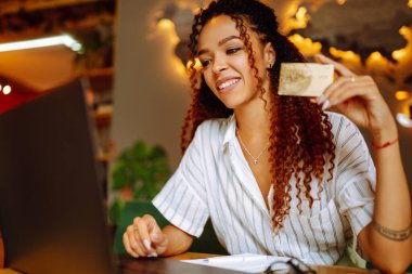 Kredi kartı tutan ve dizüstü bilgisayar kullanan genç bir kadın. Afro-Amerikalı bir kadın kafede oturmuş online alışveriş yapıyor. Çevrimiçi alışveriş, e-ticaret, internet bankacılığı, harcama.