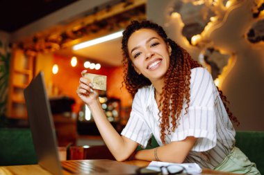 Kredi kartı tutan ve dizüstü bilgisayar kullanan genç bir kadın. Afro-Amerikalı bir kadın kafede oturmuş online alışveriş yapıyor. Çevrimiçi alışveriş, e-ticaret, internet bankacılığı, harcama.
