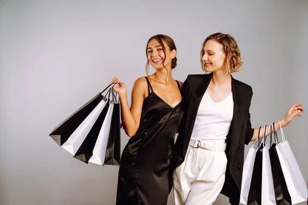 Two fashion women in black dresses with shopping bags. Young women friends with black and white bags posing on grey background. Purchases, black friday, discounts, sale concept.